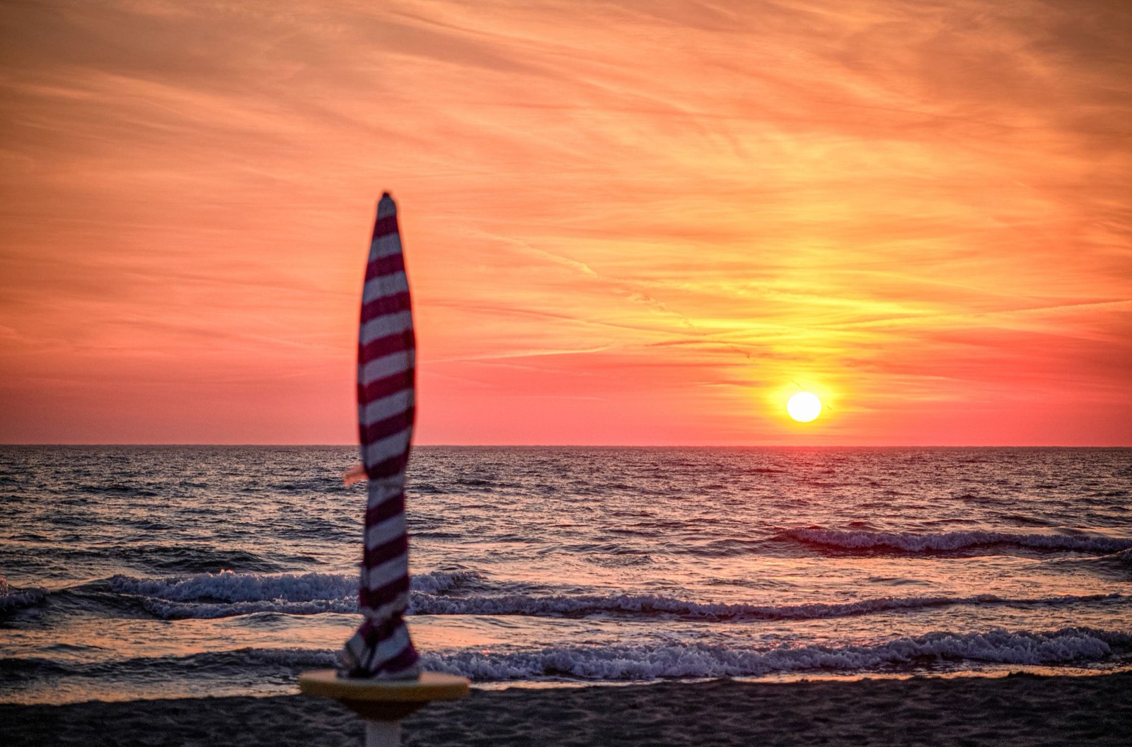 A closed striped parasol on a sandy beach at sunset, with the sun low over the sea