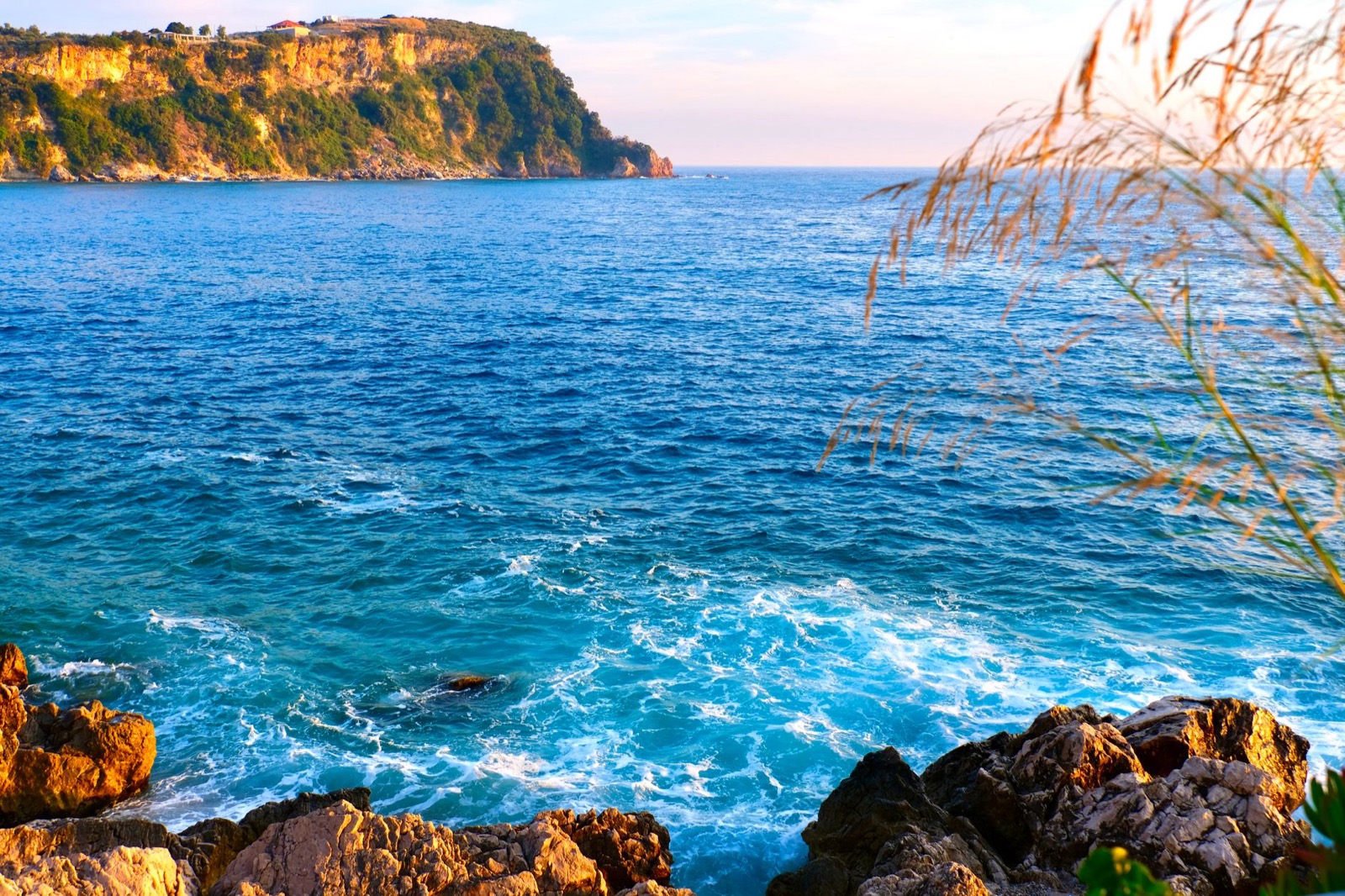 A rocky shore and bright turquoise sea below a wooded headland catching low sun