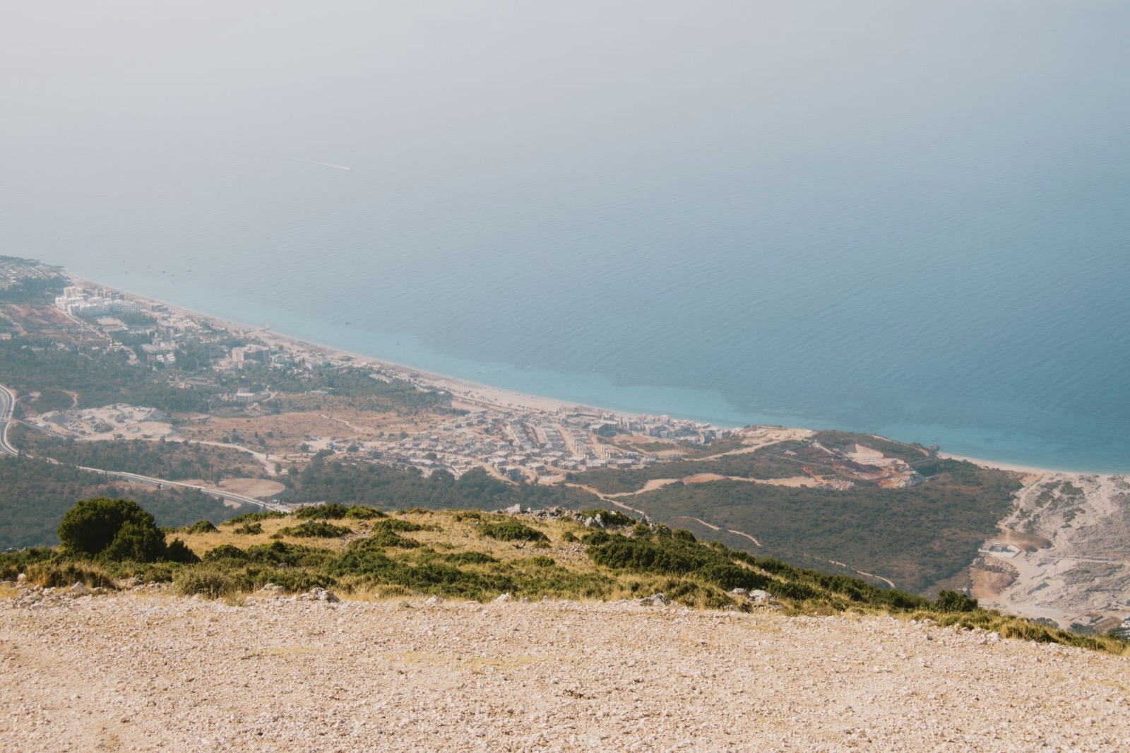 A long beach and a coastal town seen from a scrubby ridge high above a hazy sea