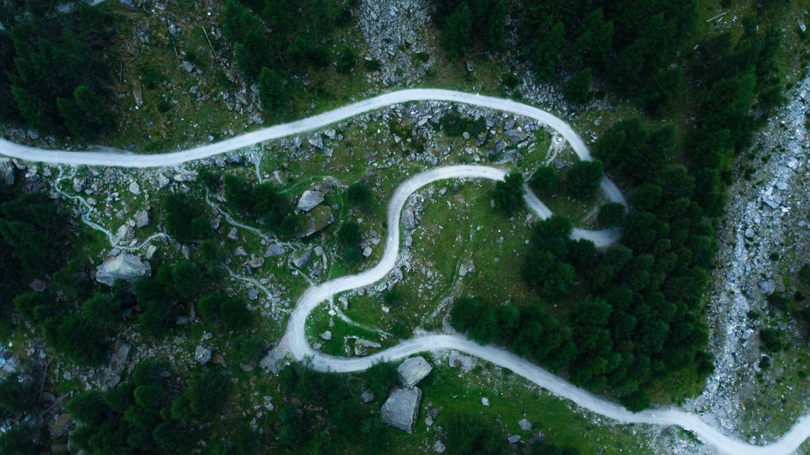 An aerial view of a narrow unsealed road in tight hairpins across a green, boulder-strewn mountainside