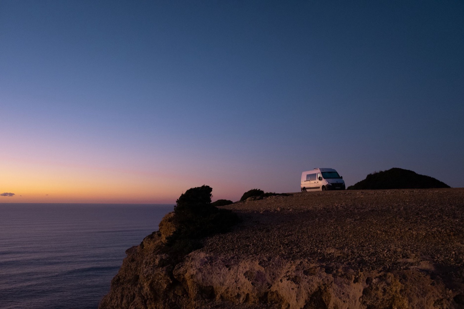 A campervan parked on a stony clifftop above the sea at dusk
