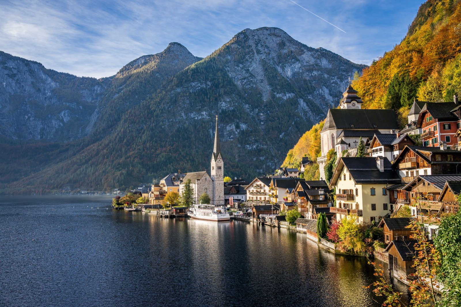 A lakeside village with a church spire below steep wooded mountains in autumn