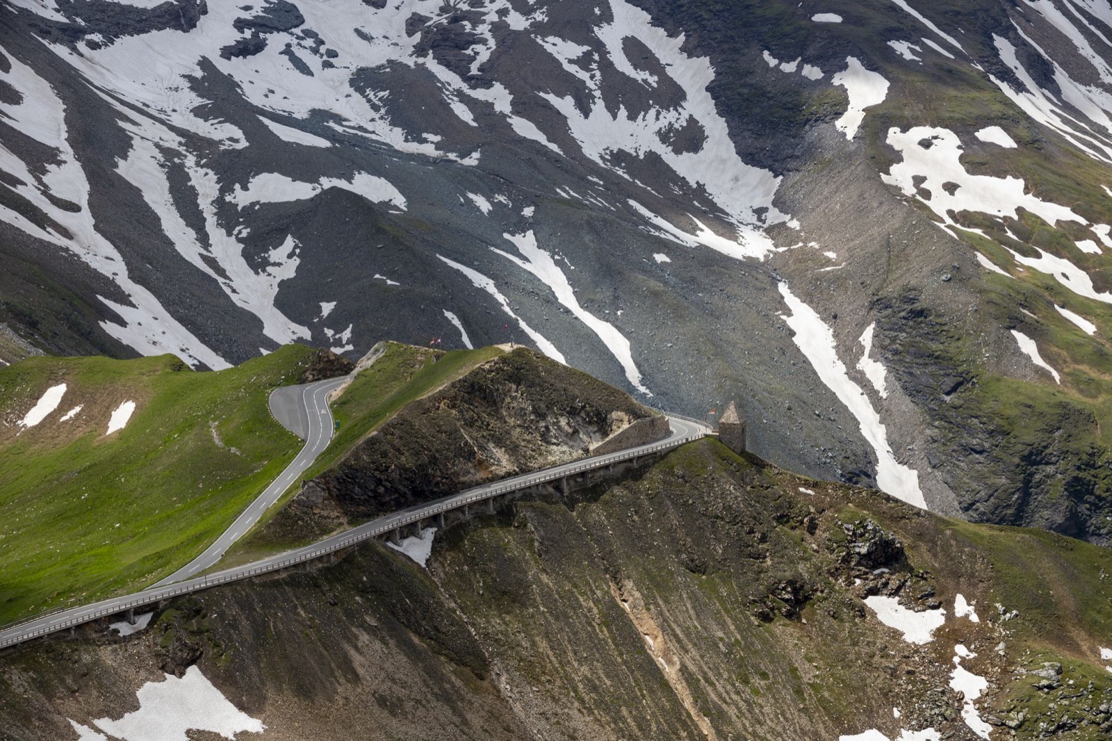 A mountain road crossing a grassy ridge on a viaduct below snow-streaked alpine slopes