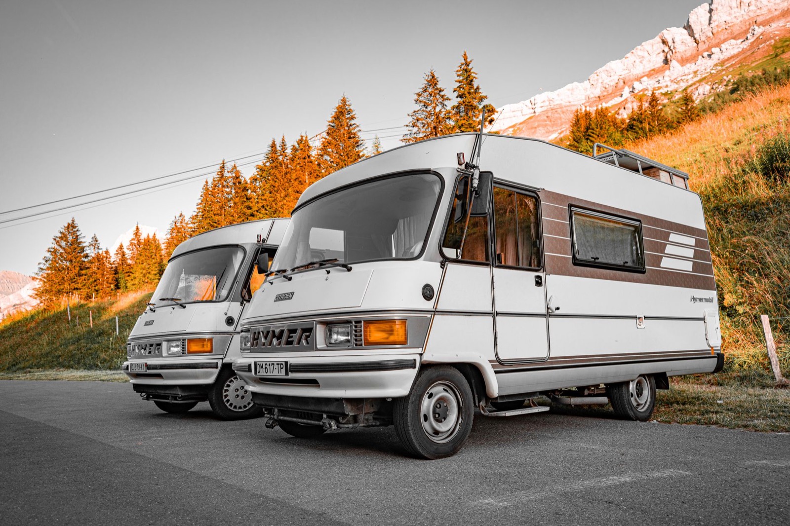 Two boxy vintage motorhomes parked on asphalt below a pine slope and a rocky peak