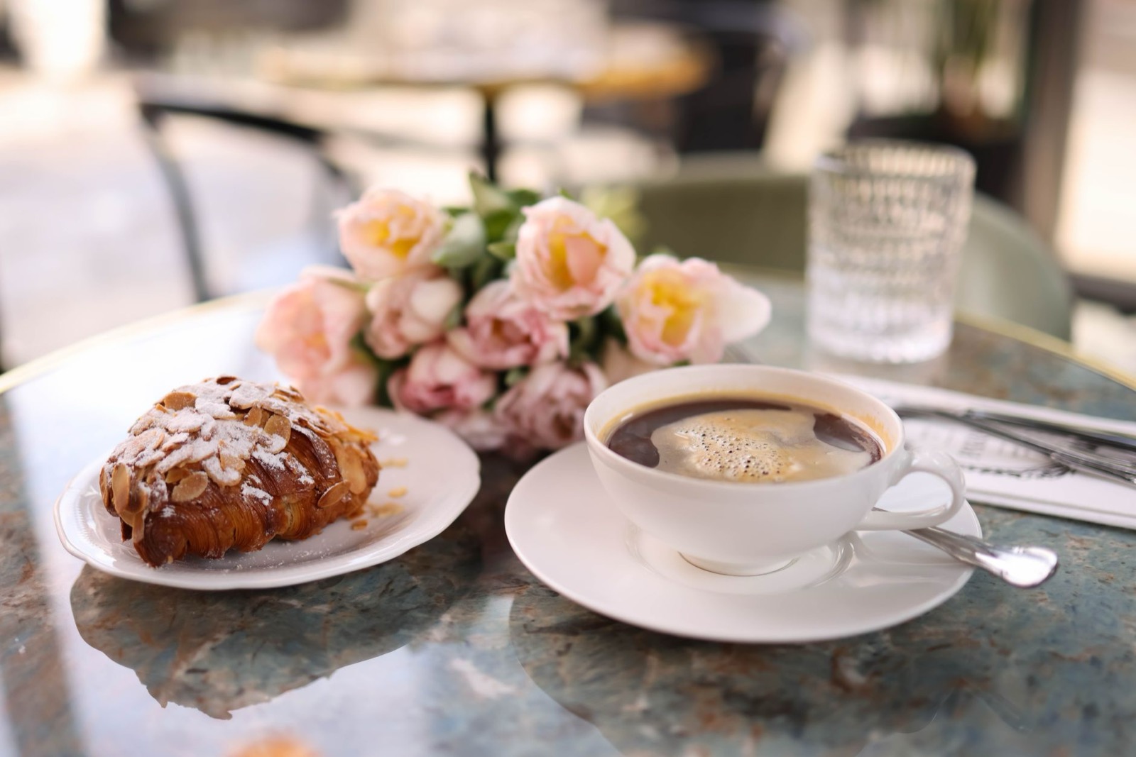 An almond croissant on a plate beside a cup of black coffee on a marble café table