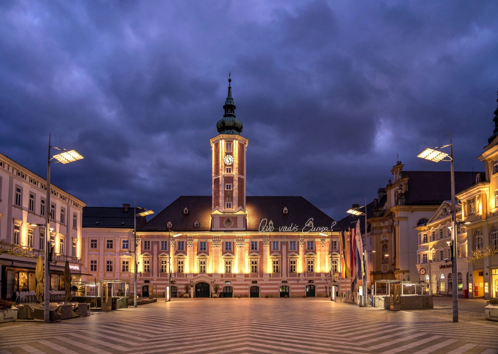A lit baroque town hall with a clock tower above a paved square at dusk