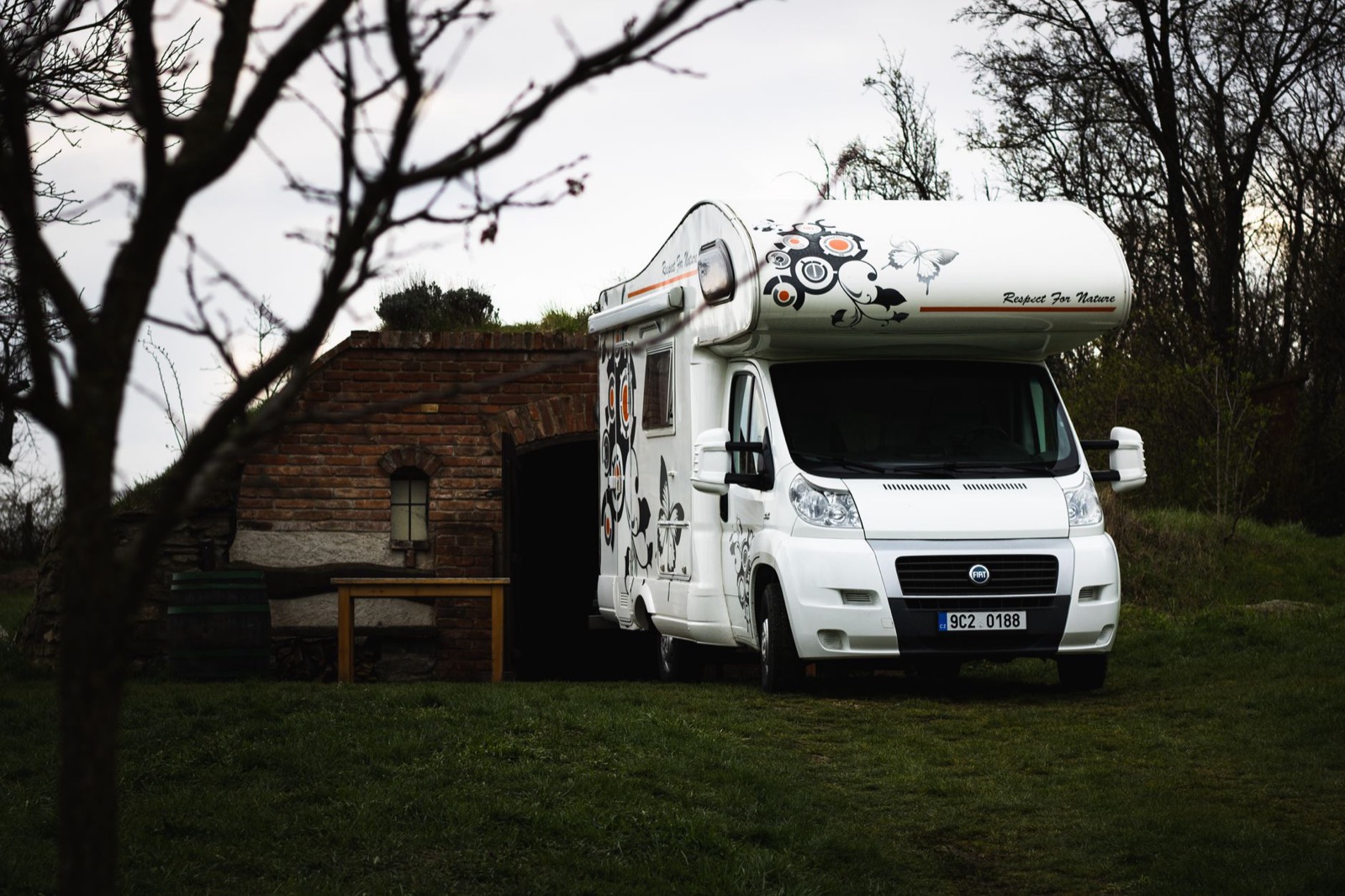A white coachbuilt motorhome parked on grass beside an old brick cellar at dusk
