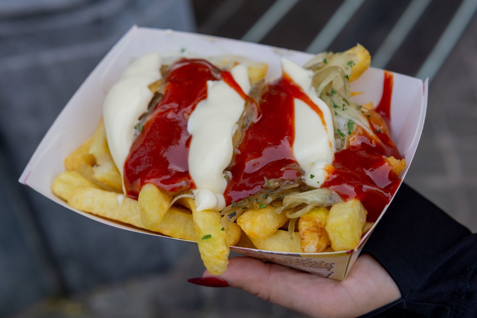 A cardboard tray of fries held in one hand, under mayonnaise, ketchup and fried onions