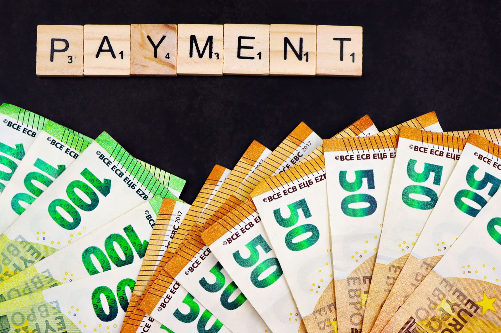 Fanned 50 and 100 euro banknotes below wooden letter tiles spelling PAYMENT