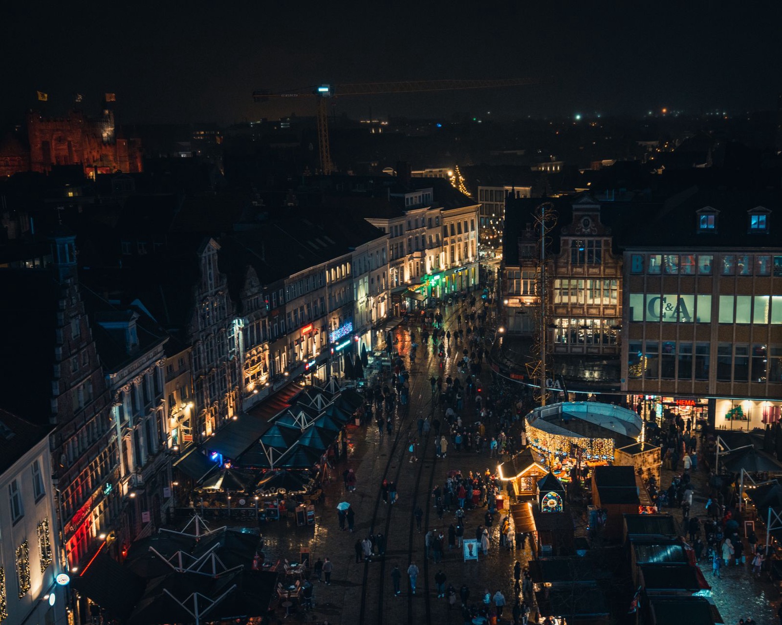 A crowded Belgian city centre at night from above, lit shopfronts and market stalls along a wet street
