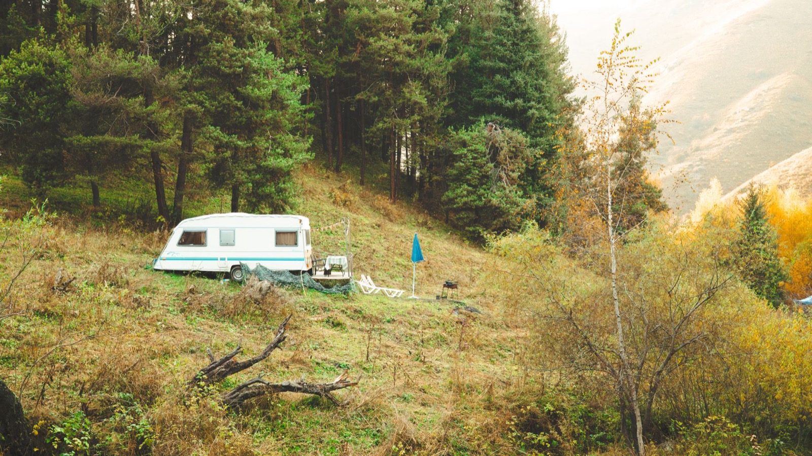 A caravan pitched on a wooded hillside with chairs, a table and a parasol set out on the grass