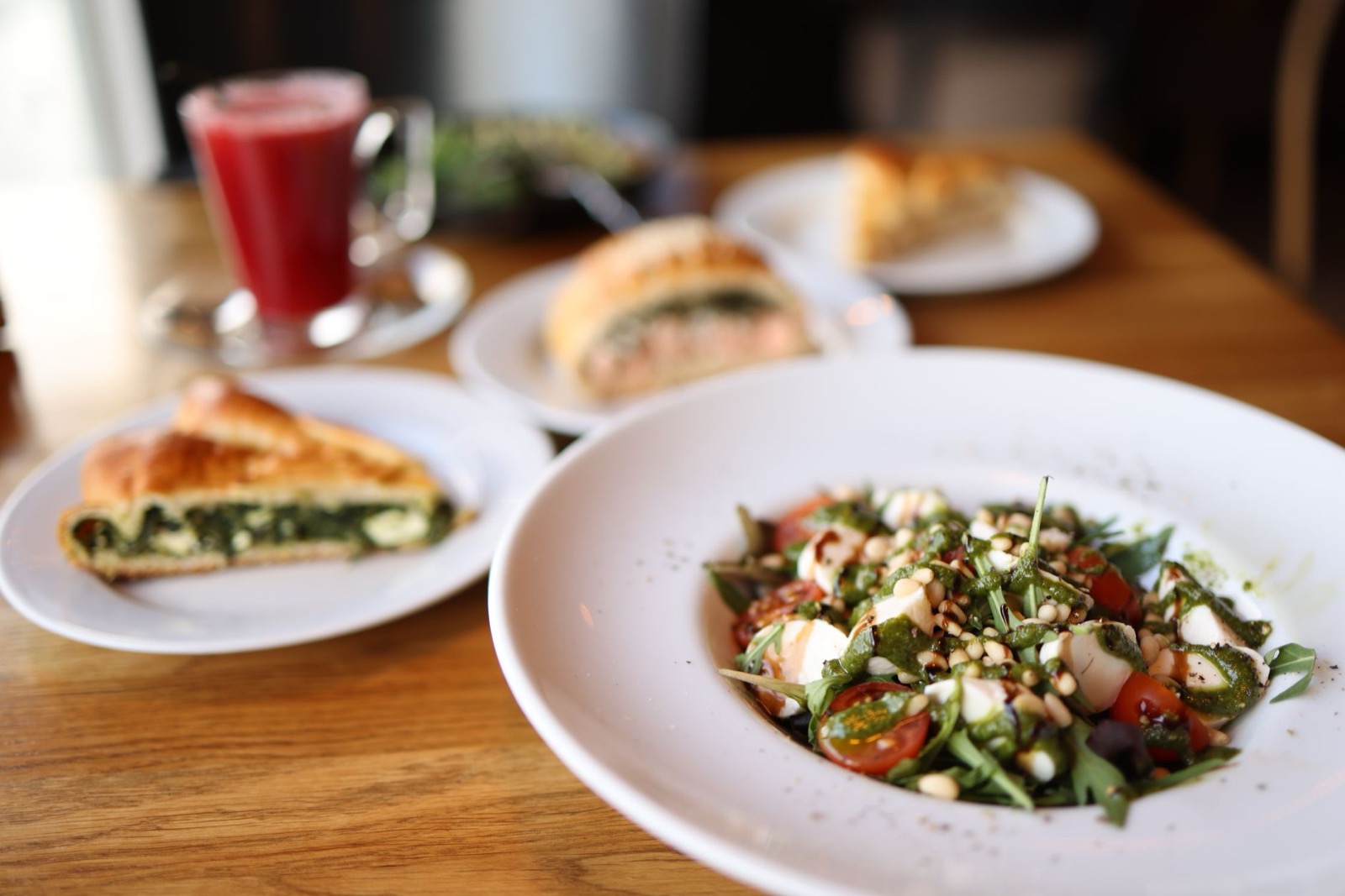A slice of spinach filled pastry beside a leaf and tomato salad on a café table, with a glass of beetroot juice