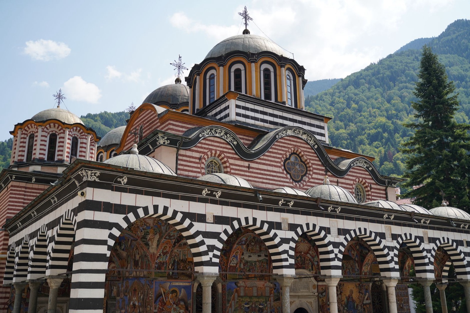 The striped arches and painted arcade of Rila Monastery below forested mountains in Bulgaria