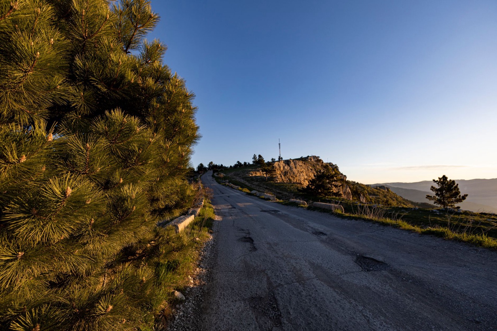 A patched and potholed asphalt road running along a pine ridge towards a rocky outcrop at sunset