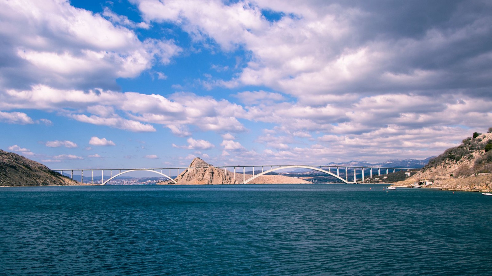 A white arch bridge carried on piers across a blue channel between bare karst hills