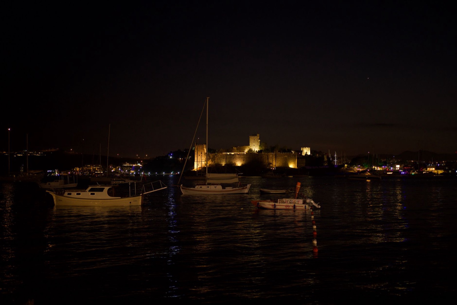 Moored boats on dark water at night with a floodlit fortress and town lights behind them