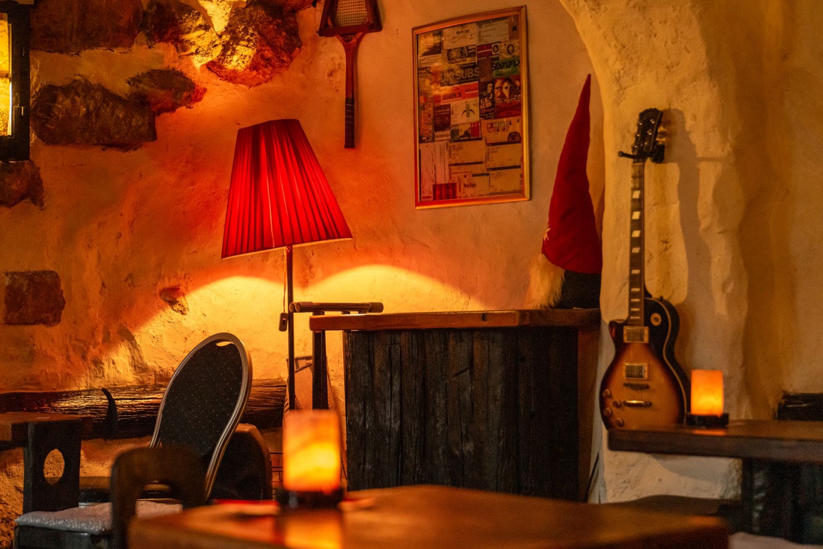 A lamplit tavern interior with rough plaster walls, wooden tables, candles and a guitar propped in the corner