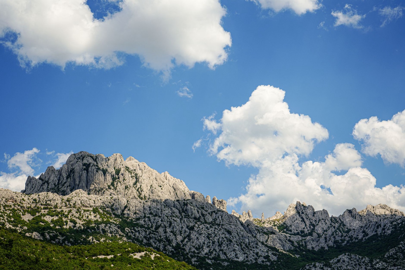 Bare grey limestone peaks rising above green wooded slopes under a bright cloudy sky