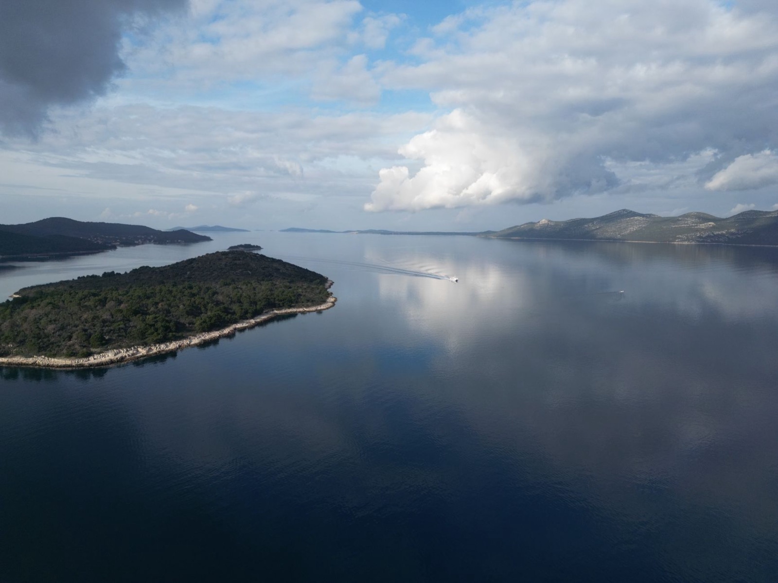 Wooded islands and headlands in a flat blue channel seen from the air, a boat leaving a wake