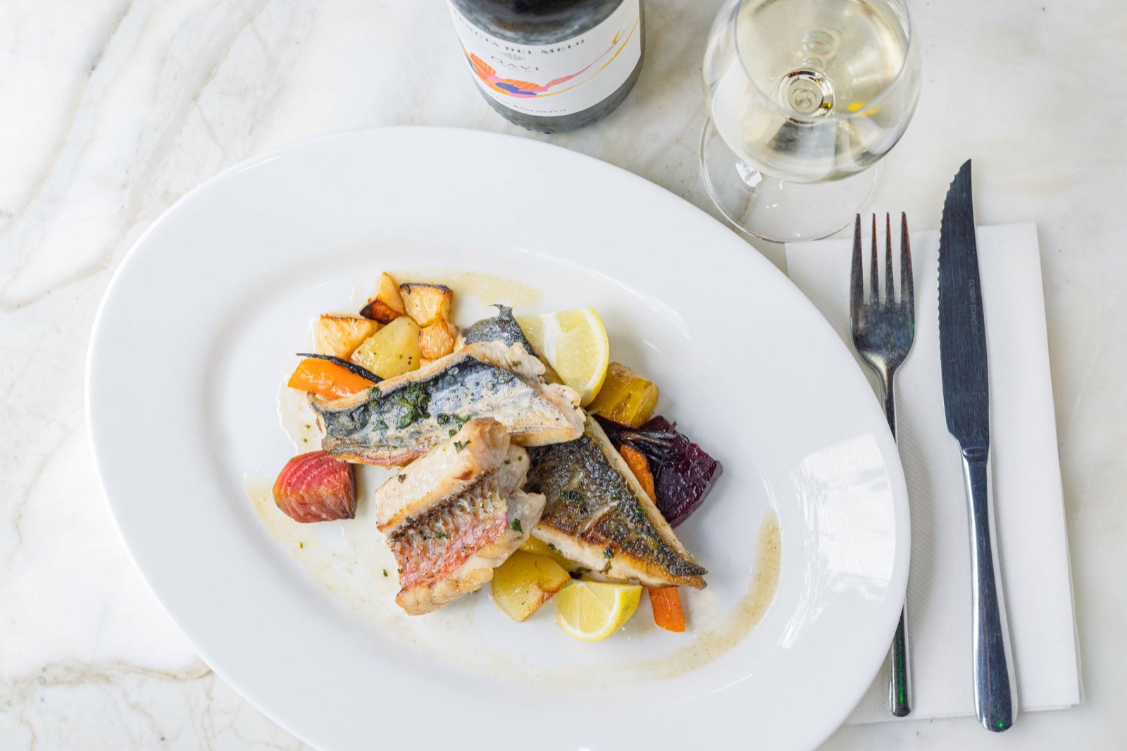 Grilled fish fillets with roast potatoes, vegetables and lemon on a white plate beside a glass of white wine