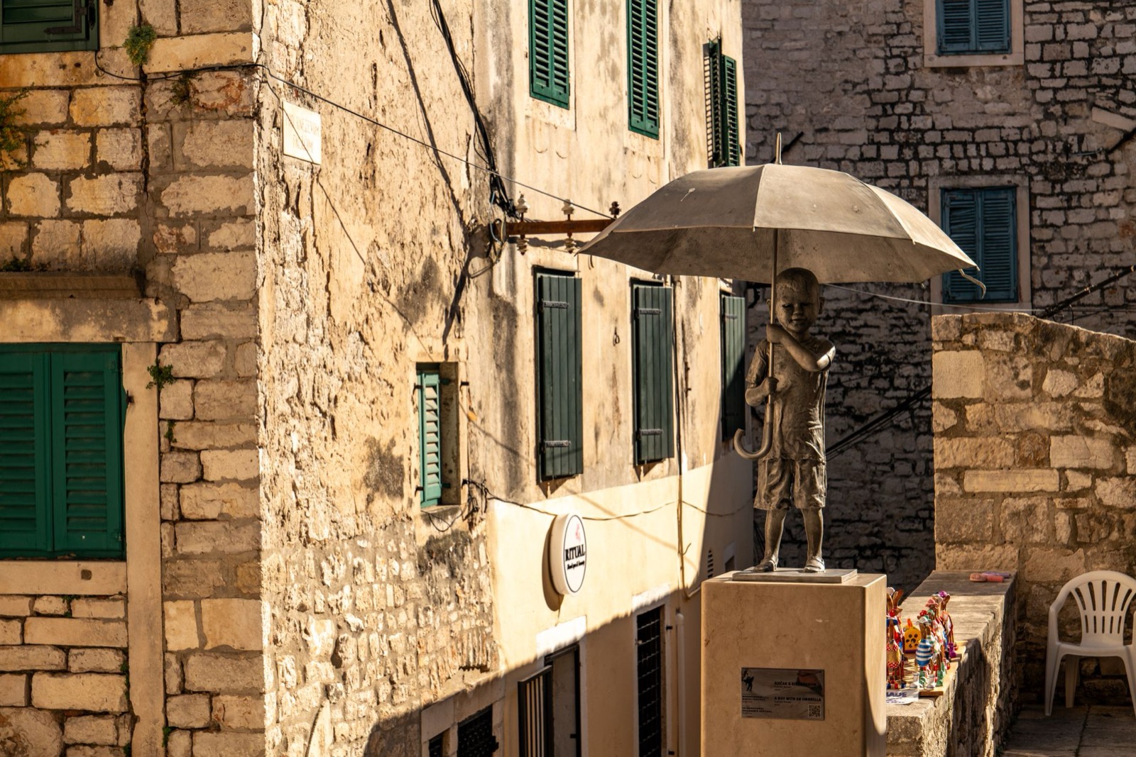 A bronze statue of a boy holding an umbrella on a corner between stone houses with green shutters