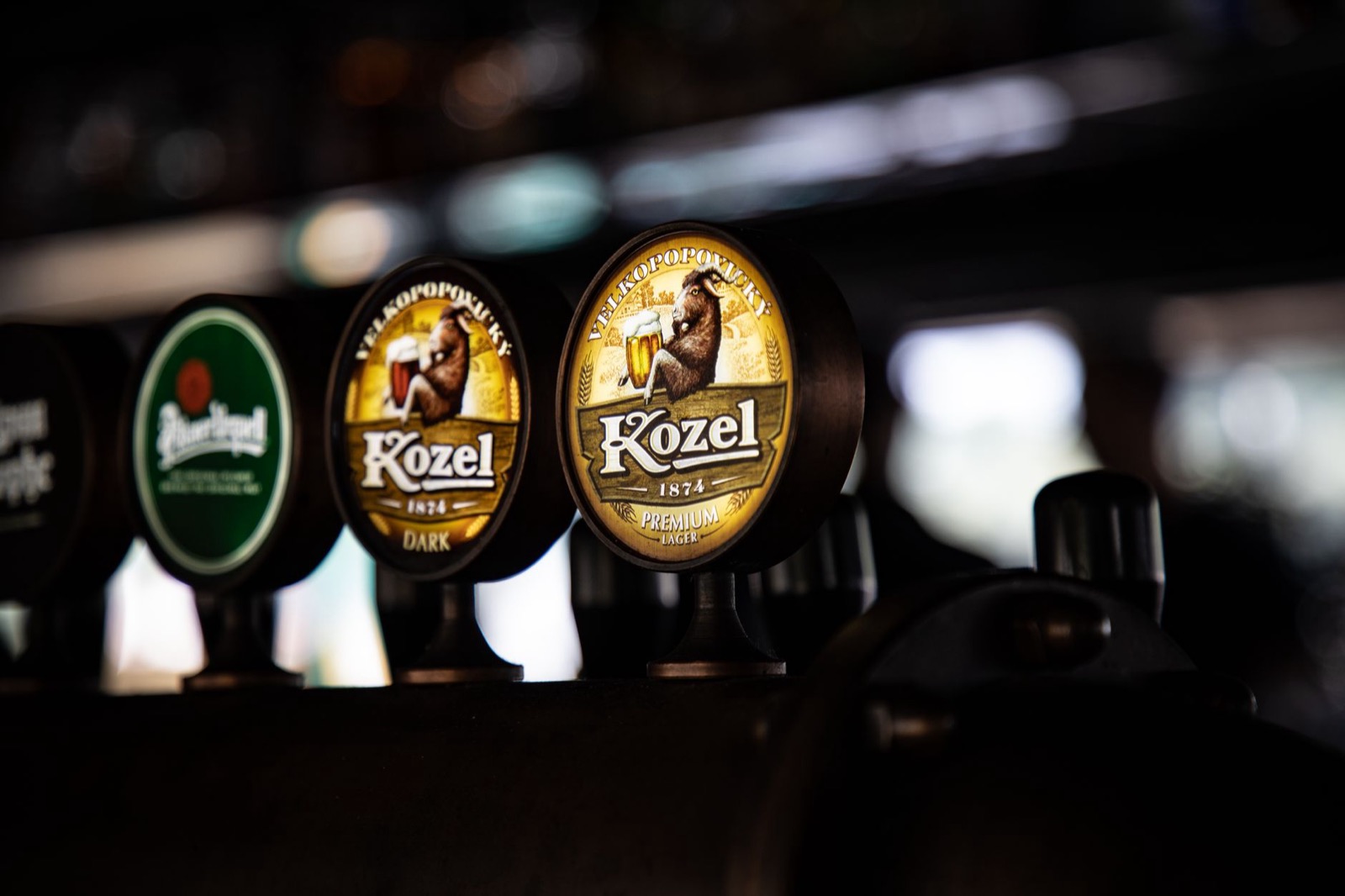 A row of lit beer tap handles on a dark bar, Kozel and Pilsner Urquell among them