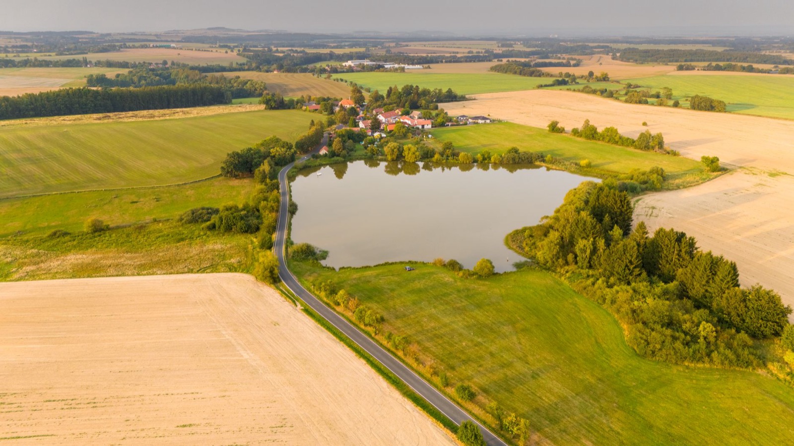 A fishpond among fields and hedgerows with a small village on its far shore, seen from the air