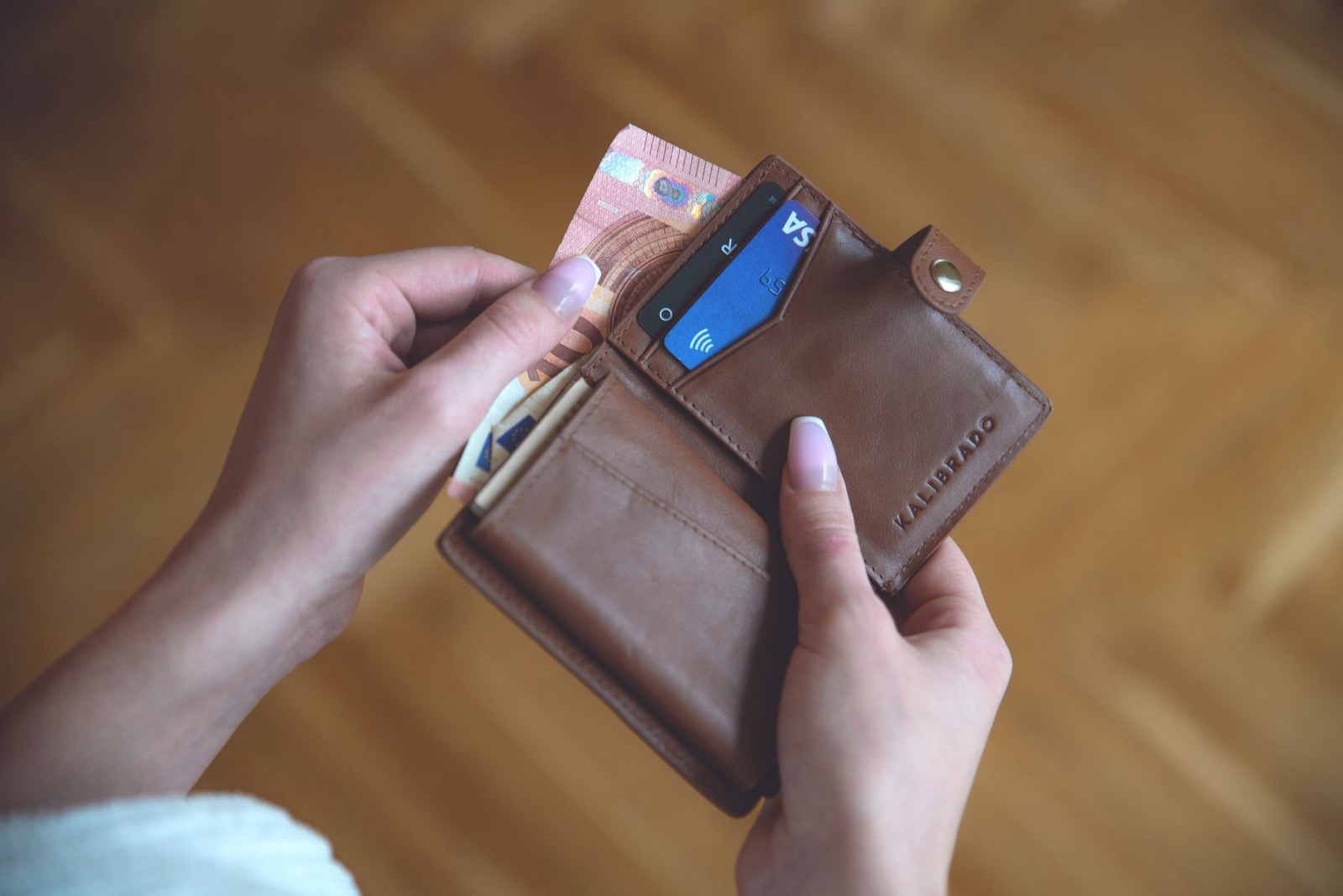 Two hands opening a brown leather wallet holding banknotes and a blue contactless card