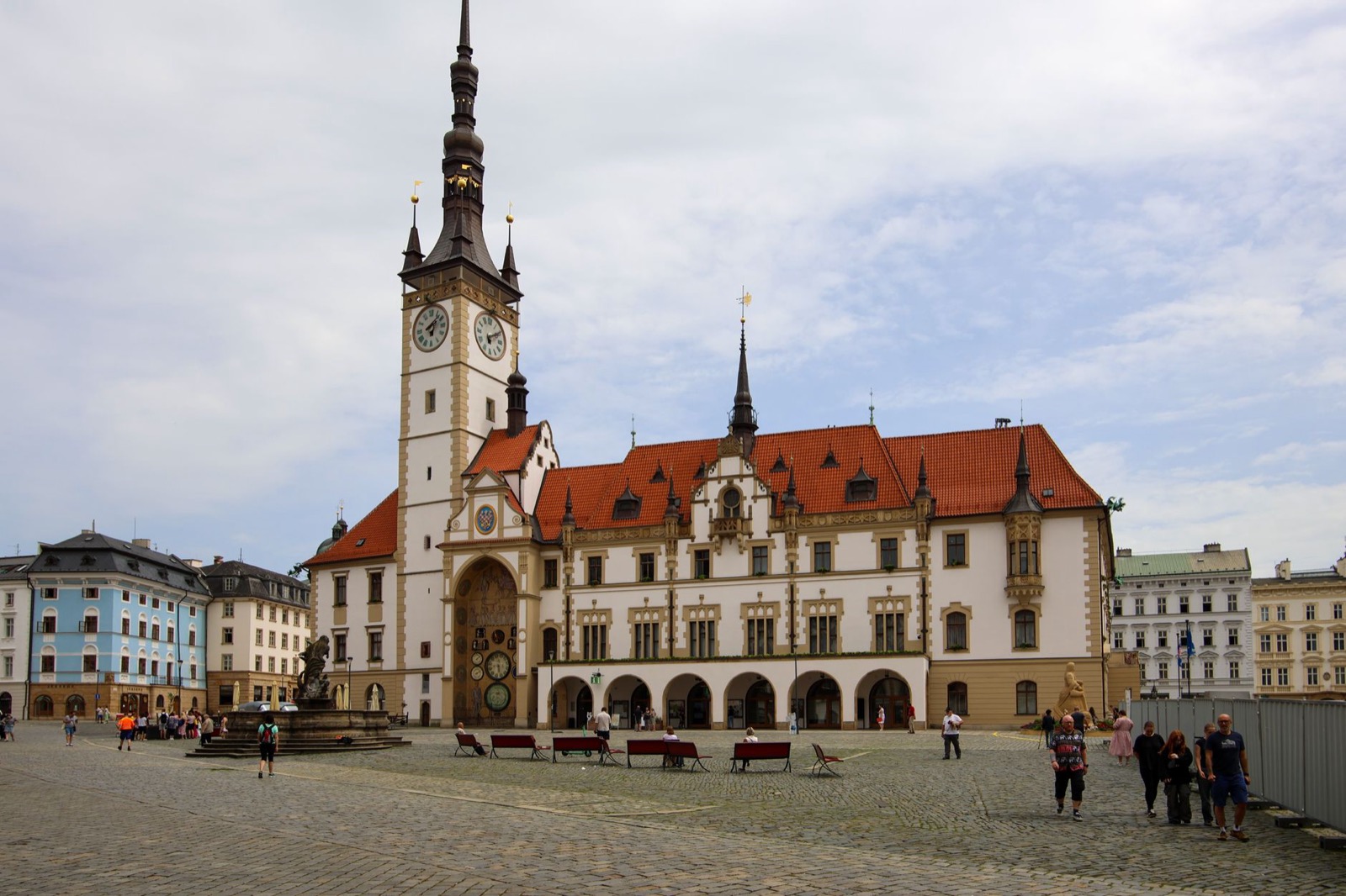 A town hall with a tall clock tower and red tiled roof across a wide cobbled square