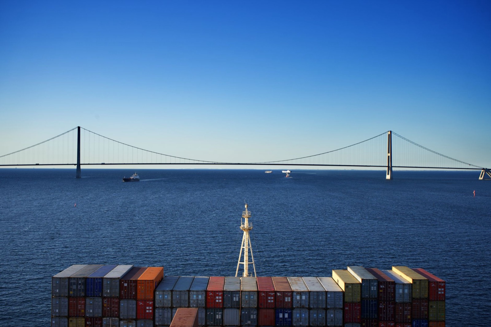 A long suspension bridge spanning open water, seen over the stacked containers of a ship