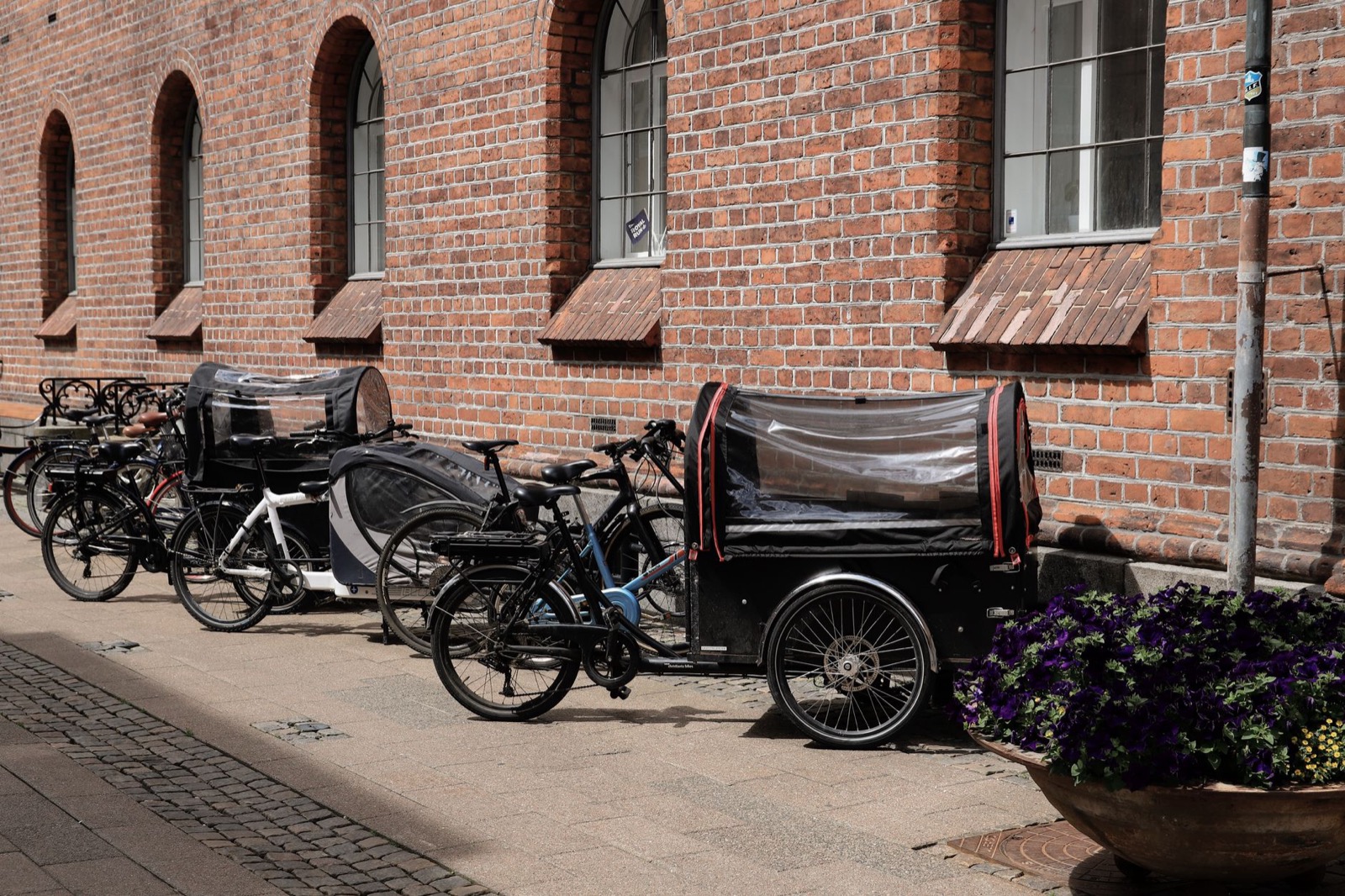 Bicycles and covered cargo bikes parked in a row along a red brick wall