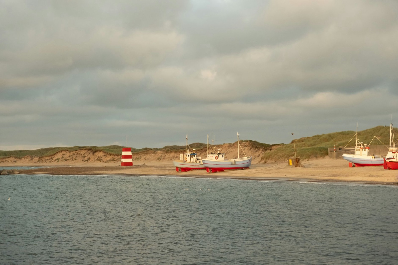 Fishing boats hauled up on the sand beside a red and white beacon below low dunes