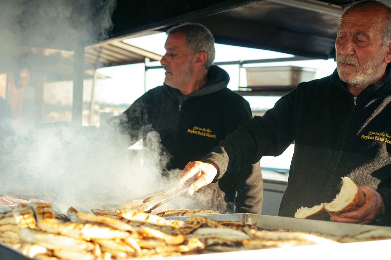 Two cooks turning grilled fish over a smoking griddle at a street food stall