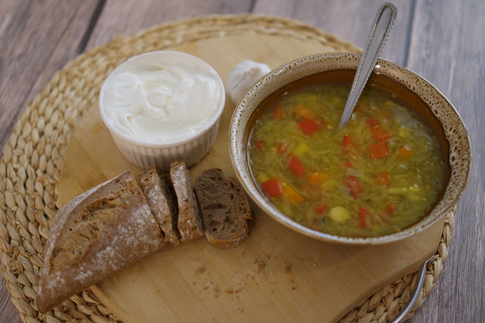 A bowl of vegetable soup with sour cream and slices of dark bread on a board