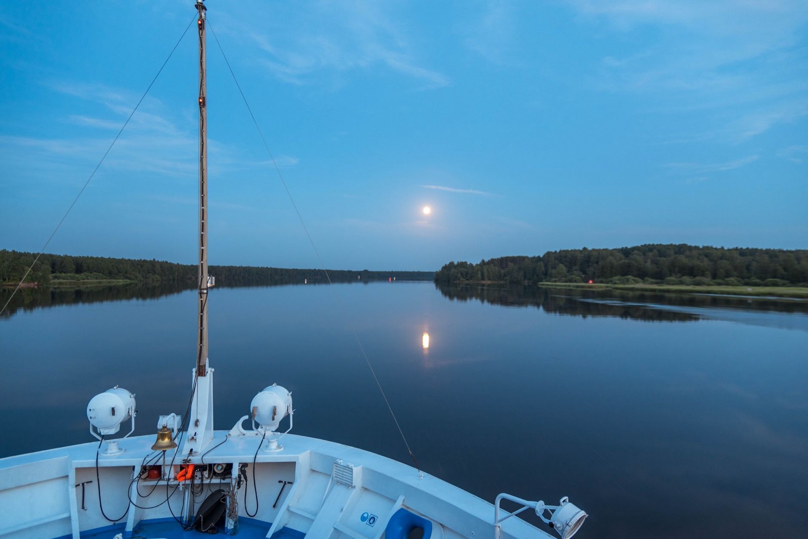 A pale sun sitting low over still water at twilight, seen over the bow of a boat