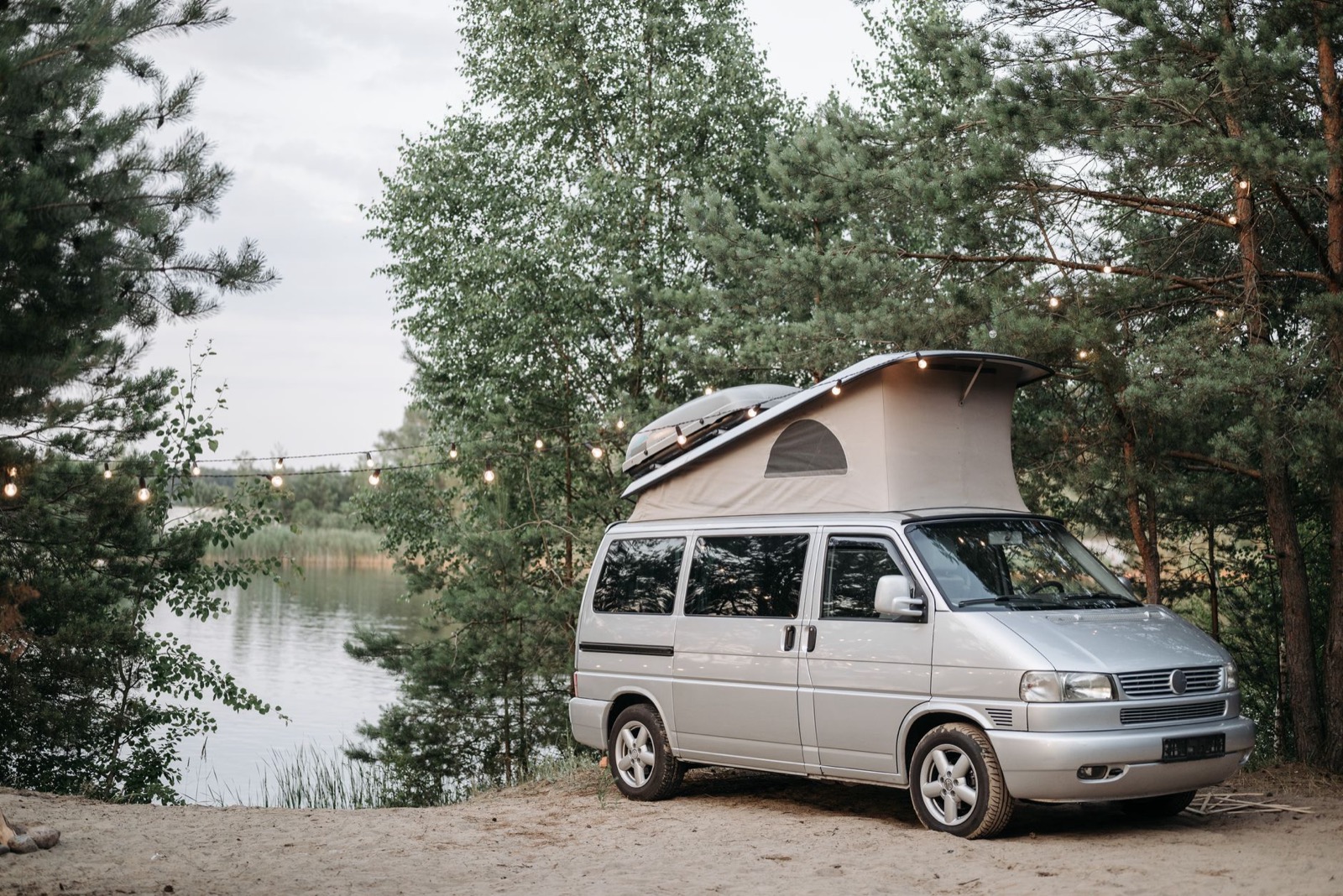 A silver pop-top camper van parked on sand beside a lake, festoon lights strung in the pines