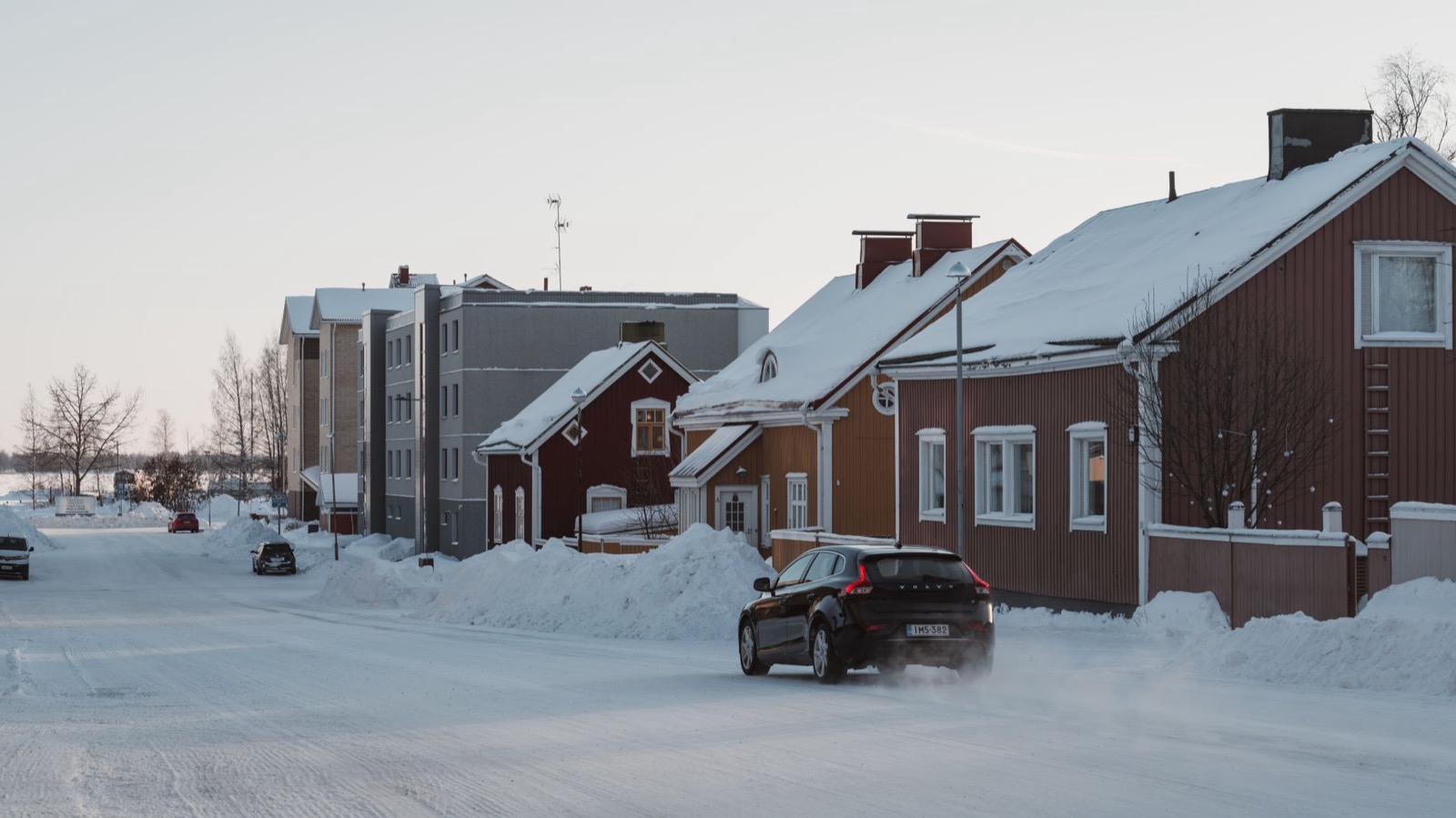A car on a snow-packed street between red wooden houses under a pale winter sky