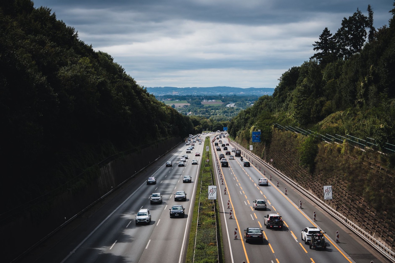 A motorway seen from a bridge, three lanes each way with steady traffic running down a wooded valley