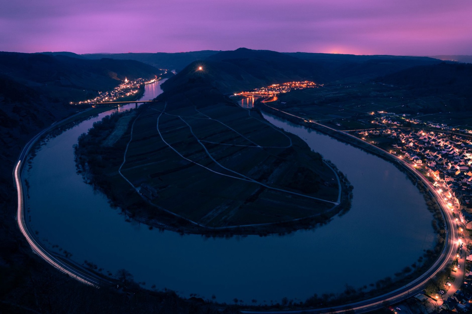 A great looping bend of the Moselle at dusk, vineyard terraces on the spur inside it and lit villages along the banks