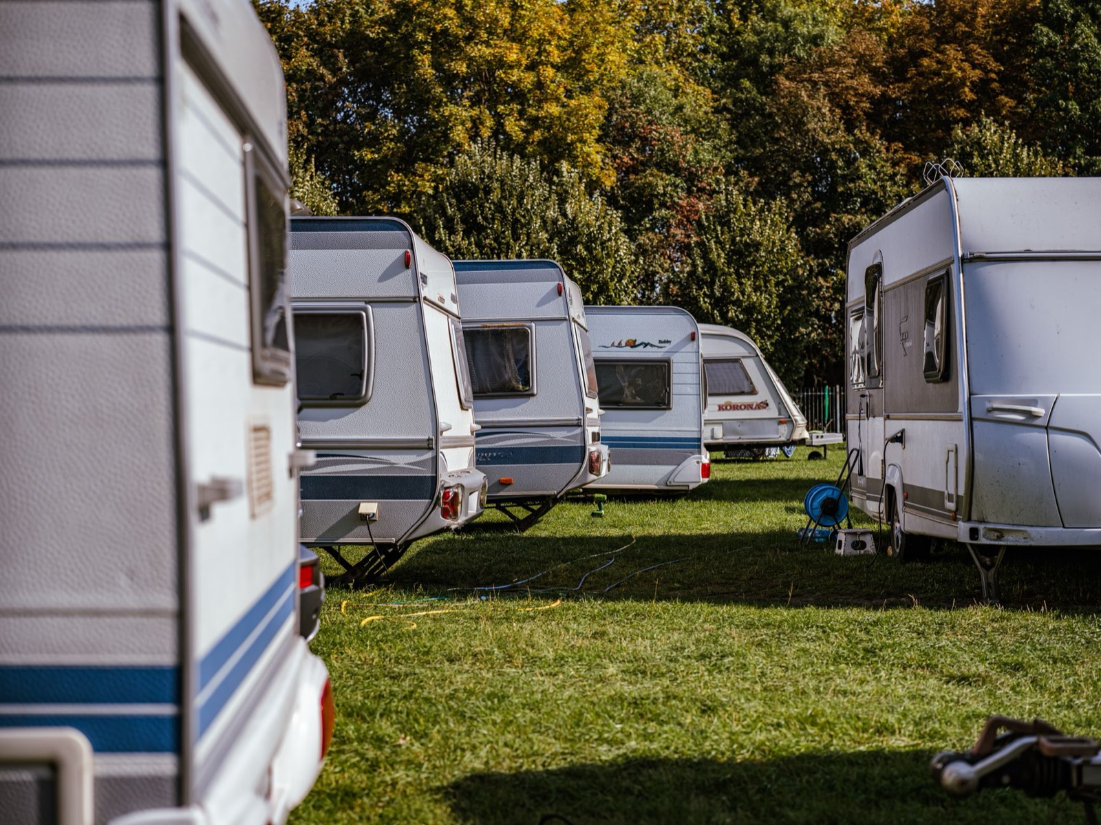 A row of white touring caravans parked on grass under autumn trees, a hose reel and a power box standing between them