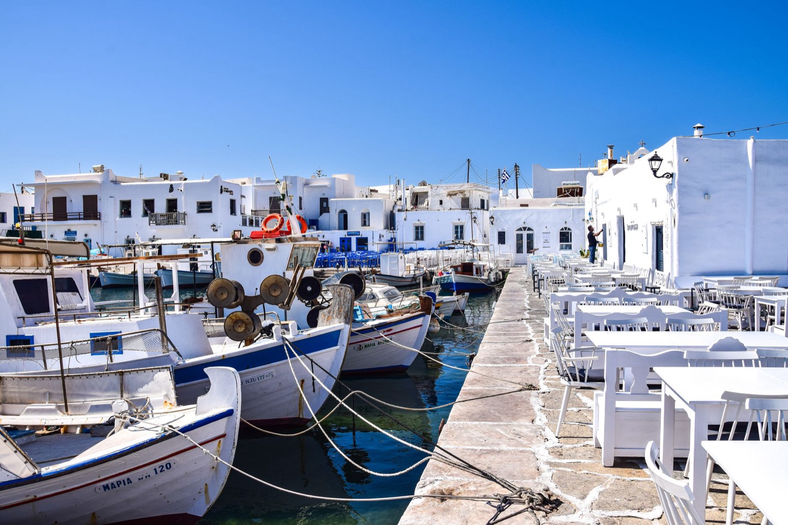 Fishing boats moored along a whitewashed Greek harbour with taverna tables set out on the quay