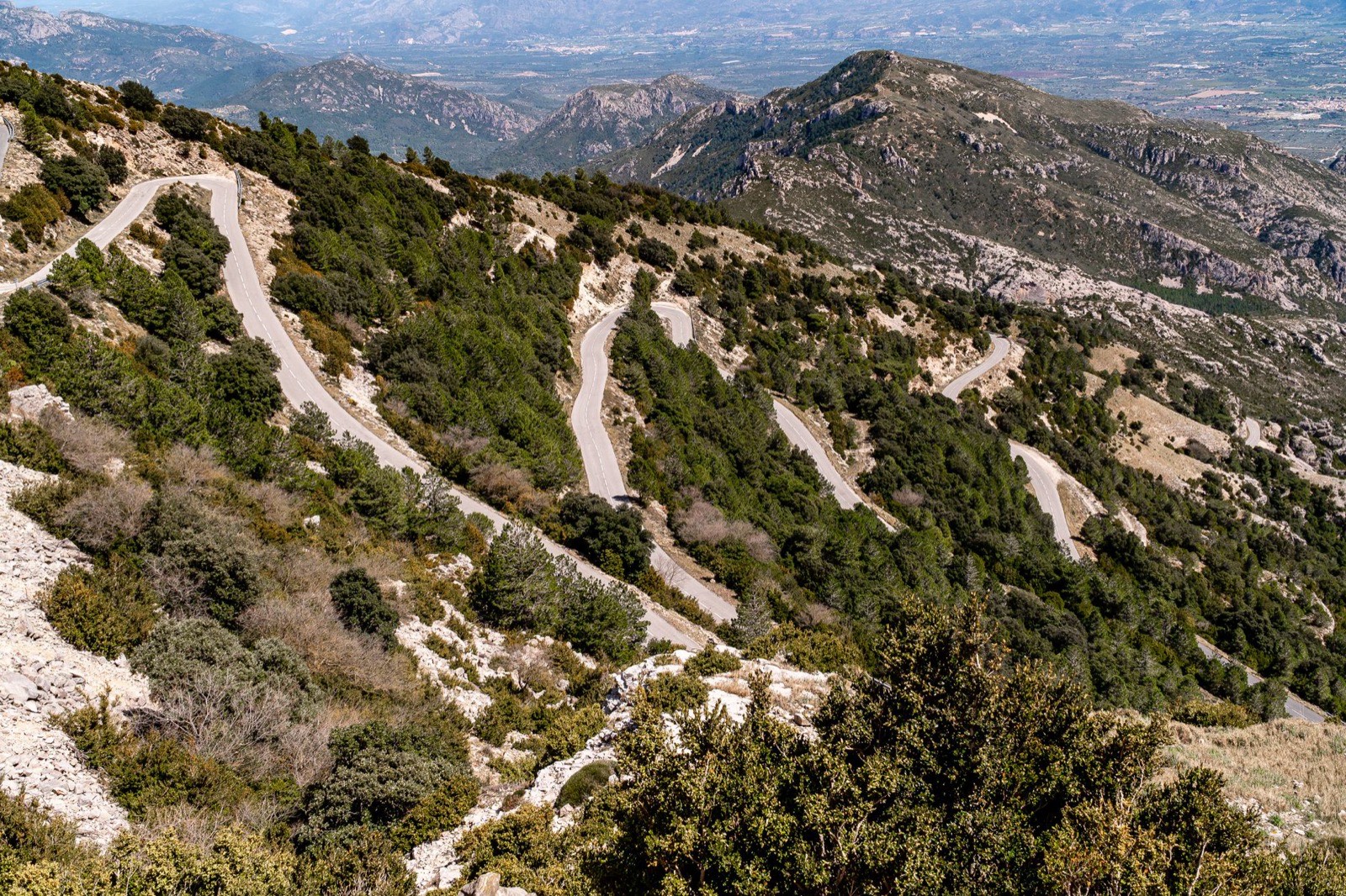 A road switchbacking down a scrubby limestone mountainside above a wide valley