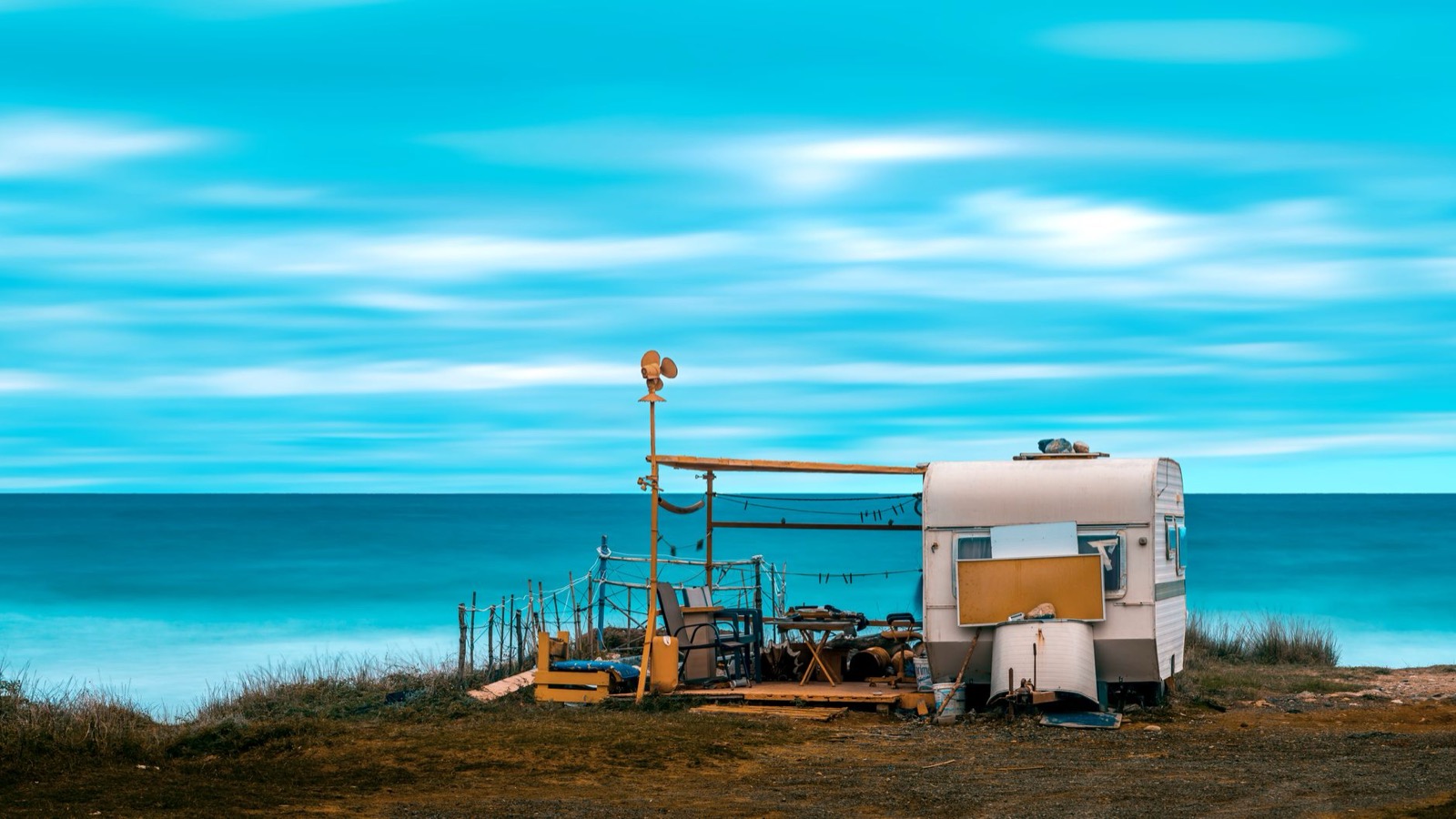 An old caravan on a grassy bluff above a bright blue sea, with tables and frames set up around it