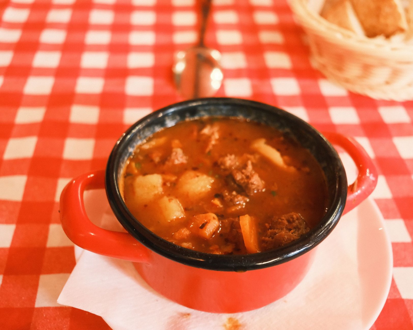 A small red enamel pot of goulash soup with beef and potato, on a red checked cloth beside a bread basket