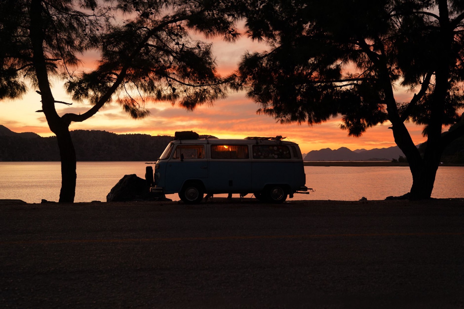 A campervan silhouetted under pine trees beside still water at sunset