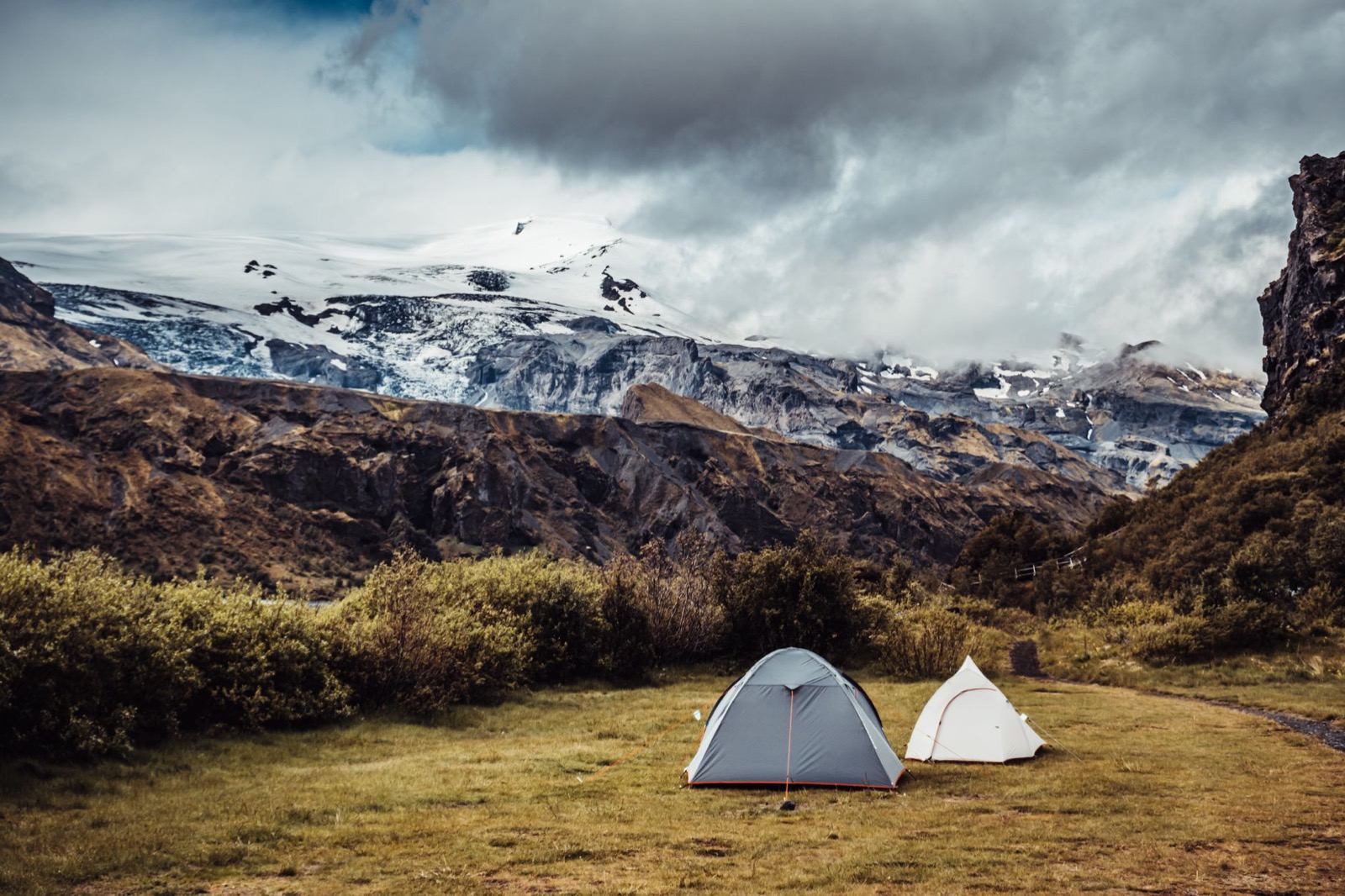 Two small tents pitched on grass below a glacier-capped mountain under heavy cloud