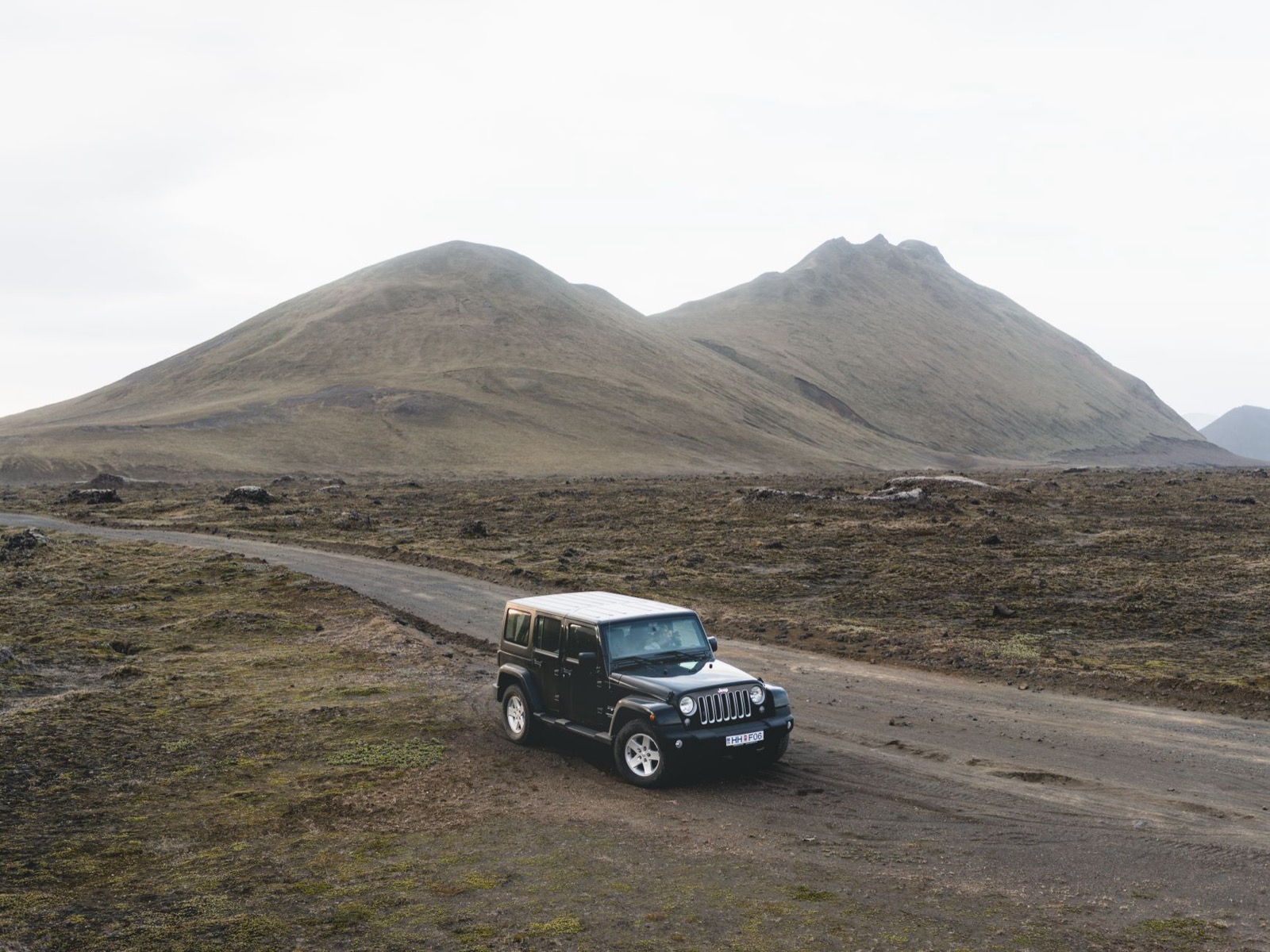 A black four wheel drive on a gravel track across bare volcanic ground below smooth brown hills