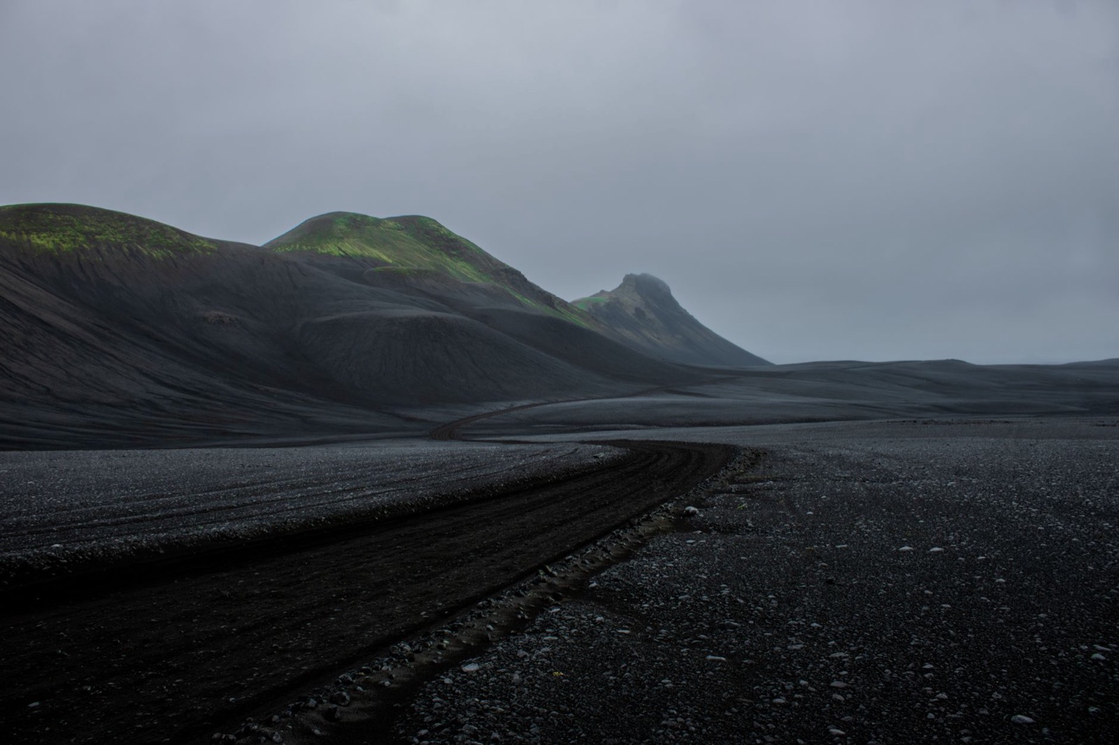 A track curving across black volcanic sand toward dark moss-streaked hills under low cloud