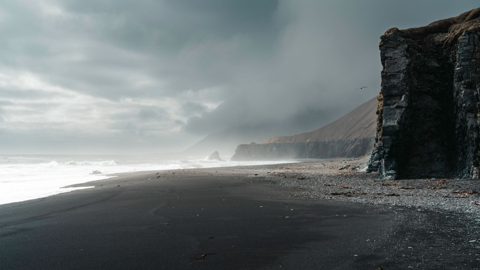 Low cloud over a black sand beach below dark cliffs, surf breaking along the shore