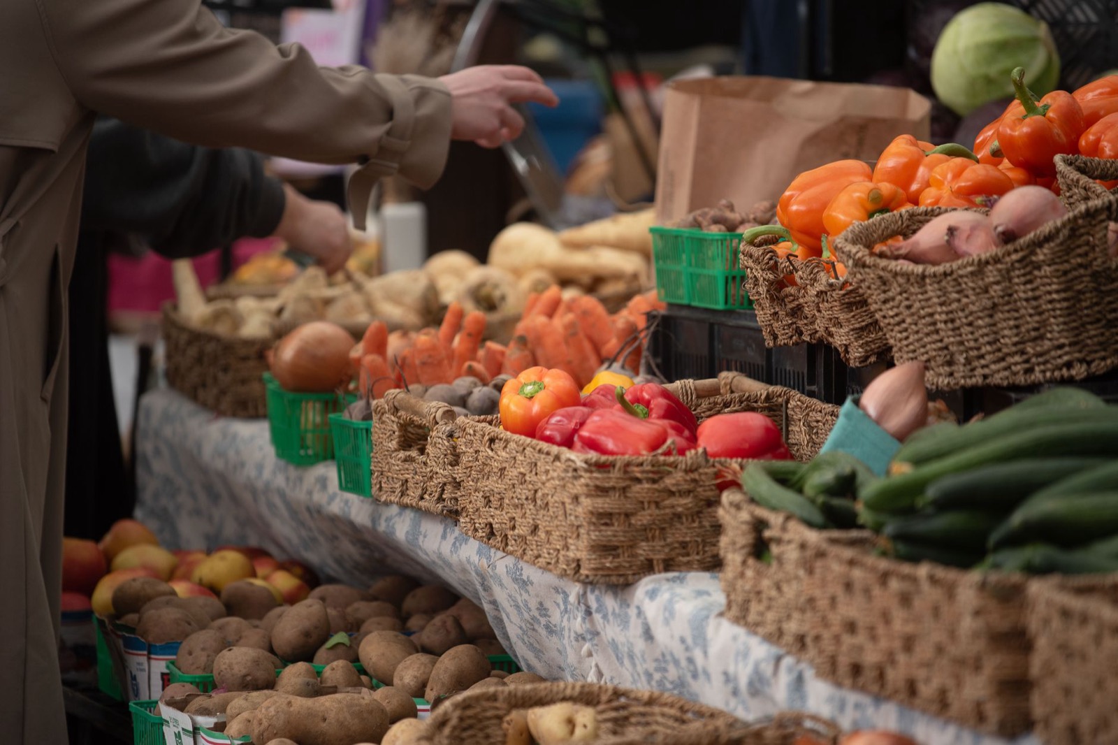 Wicker baskets of peppers, carrots and potatoes on a market stall with a customer reaching across